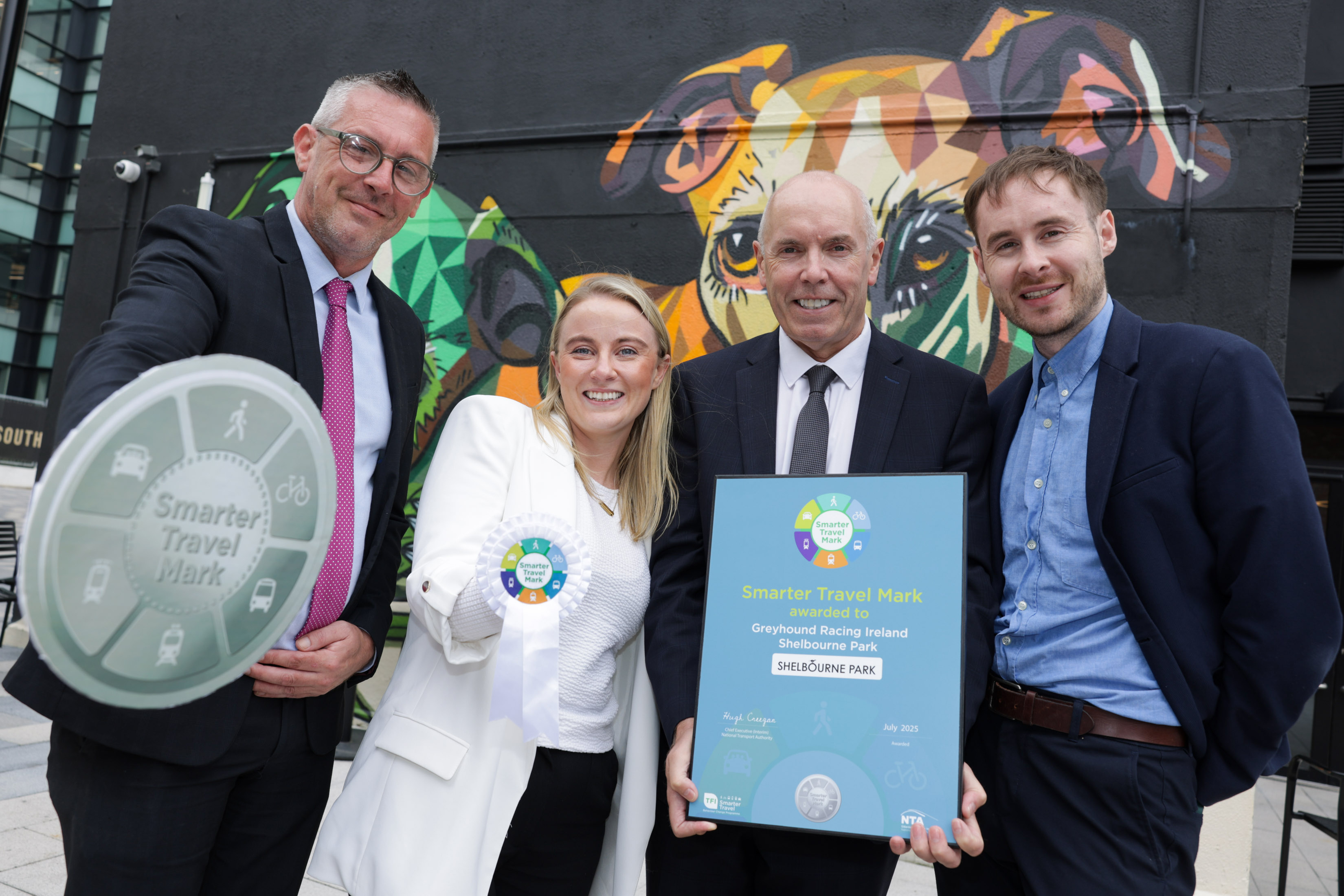 Image shows four people,  Thomas Ryan (RCÉ),  Louise Culliney of the National Transport Authority (NTA), Tim Lucy of Rásaíocht Con Éireann (RCÉ) and Shane Hayes of the National Transport Authority (NTA)., standing against a colourful mural at Shelbourne Park Greyhound Stadium to announce the Smarter Travel Award for Shelbourne Park Greyhound Stadium