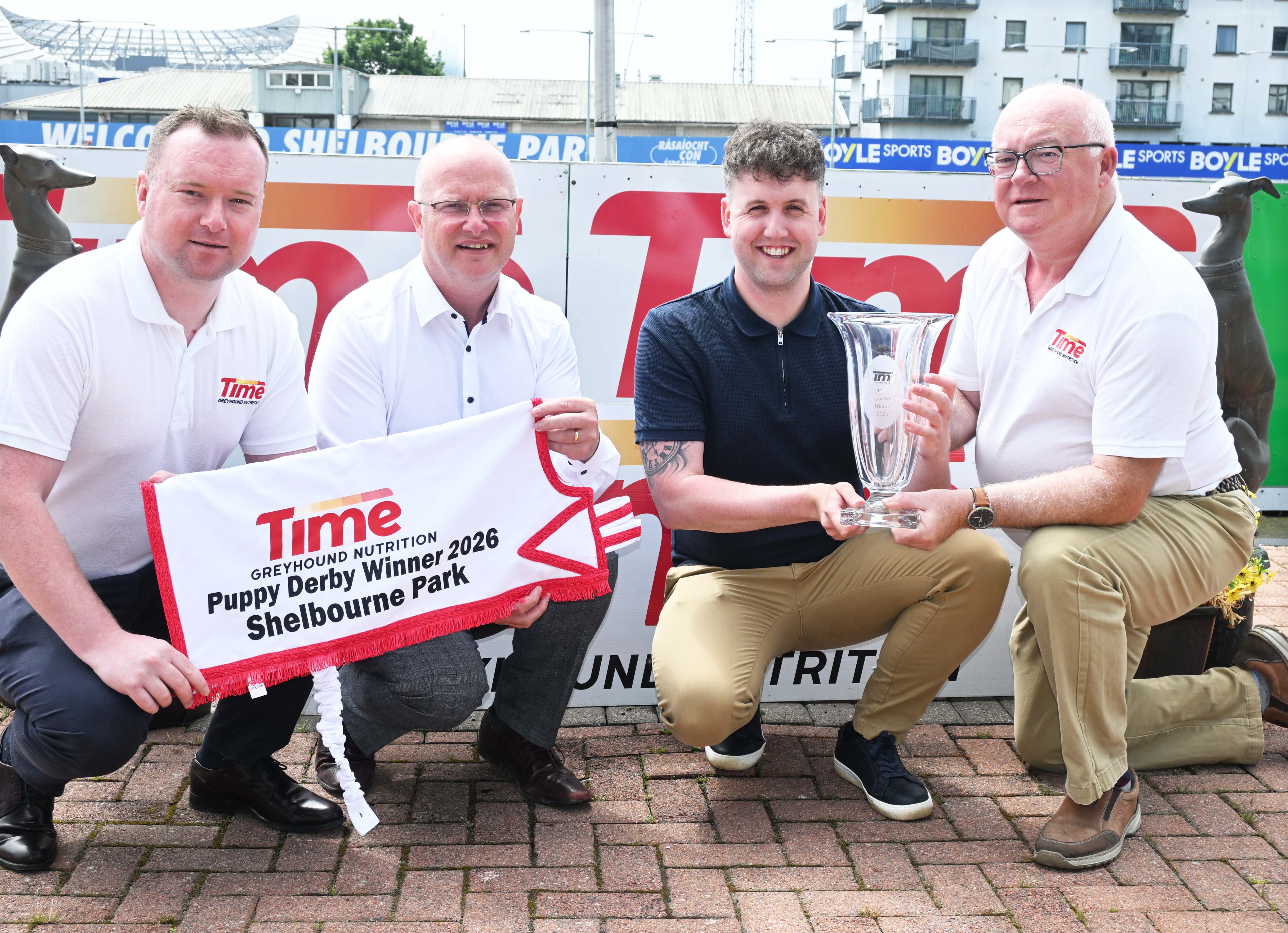 The Time Puppy Derby takes place this July in Shelbourne Park,. Sponsors WIllie Rigney and Shane Rigney of Time are pictured with the racing team in Shelbourne Park to launch the event Image shows four men pictured at Shelbourne Park Greyhound Stadium on a sunny day, holding a greyhound presentation sheet to laucn the Time Puppy Derby